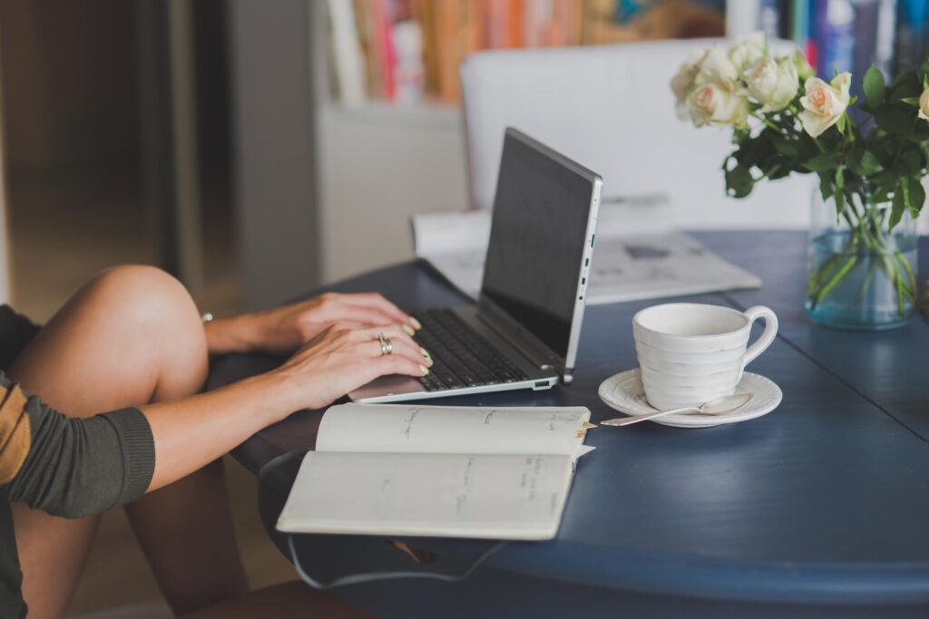 Woman typing on the laptop, coffee, notebook, and flowers on the desk next to it
